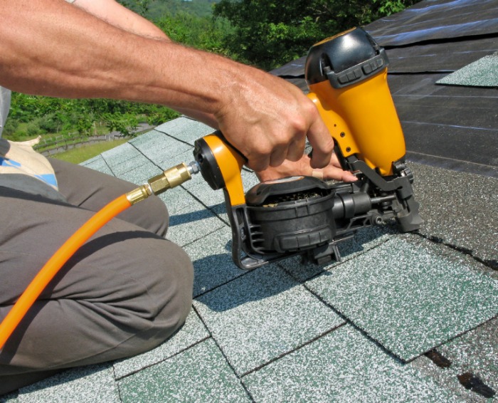 A roofing contractor uses a nail gun on the asphalt shingles. The shingles are a familiar shape, a 3-tab or strip shingles, and in a sage color.